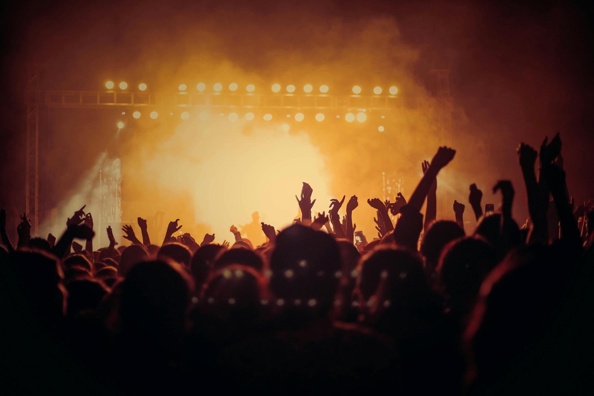 People in the crowd of a concert with event insurance, hands in the air looking toward an orange lit up stage