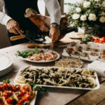 Food station at an event with various bit sized snacks to choose from and a chef carving a ham leg