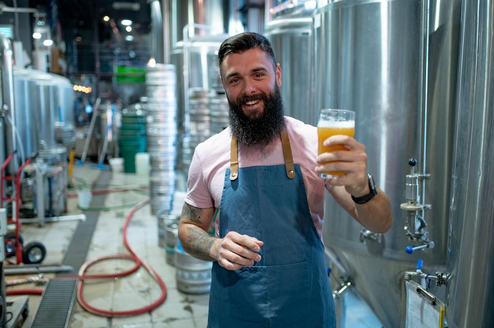 Christmas party ideas in brisbane include doing a brewery tour. Bearded man holding up a pint of beer in front of the brewing equipment