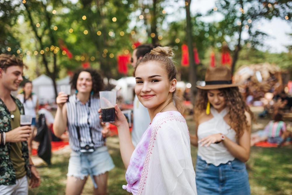 A young woman looking at the camera and smiling while holding a drink and enjoying outdoor events with a group of friends