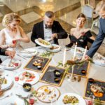 Elegently dressed banquet attendees around a table covered with food and drinks who may want to know how to plan a banquet