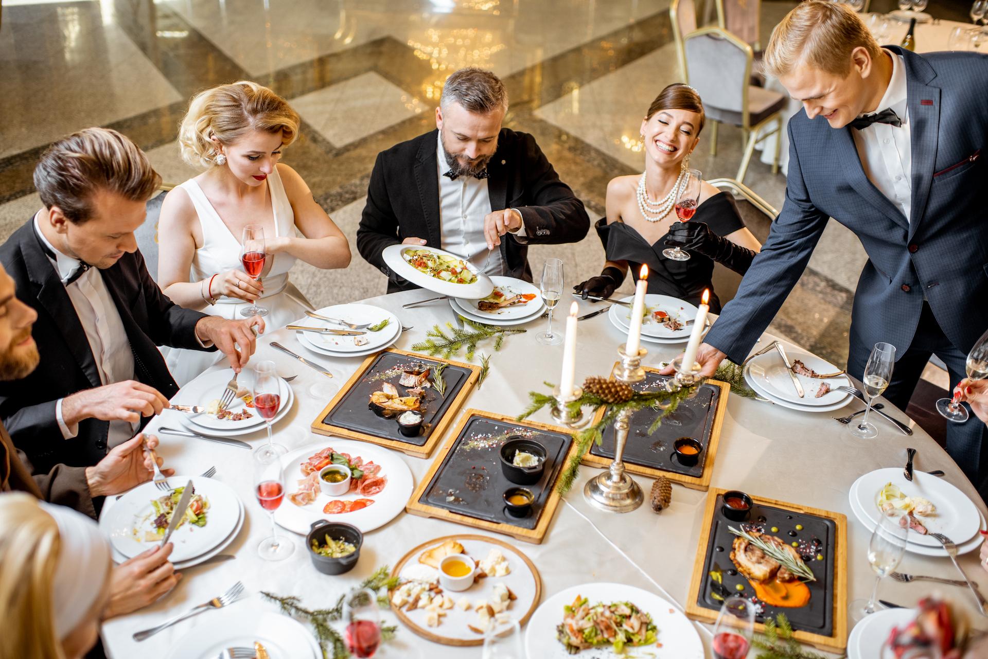 Elegently dressed banquet attendees around a table covered with food and drinks who may want to know how to plan a banquet