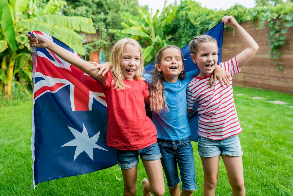 Three young girls looking happy at an Australia Day Event, flying an Australian flag behind their backs